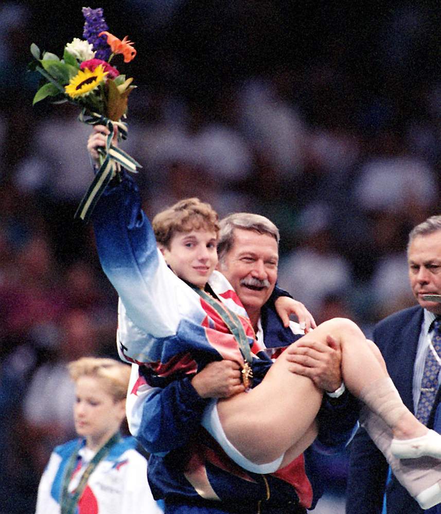 Kerri Strug hoists her flowers to the cheering crowd as she is carried from the podium by team coach, Bela Karolyi at the Georgia Dome on Tuesday, July 23, 1996. (Skip Peterson\/AJC) Kerri Strug hoists her flowers to the cheering crowd as she is carried from the podium by team coach, Bela Karolyi at the Georgia Dome on Tuesday, July 23, 1996. (Skip Peterson\/AJC)